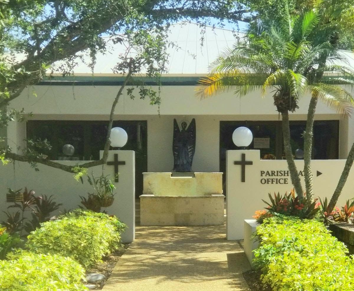Exterior view of the parish office building surrounded by greenery and decorative plants, featuring a statue and a cross.