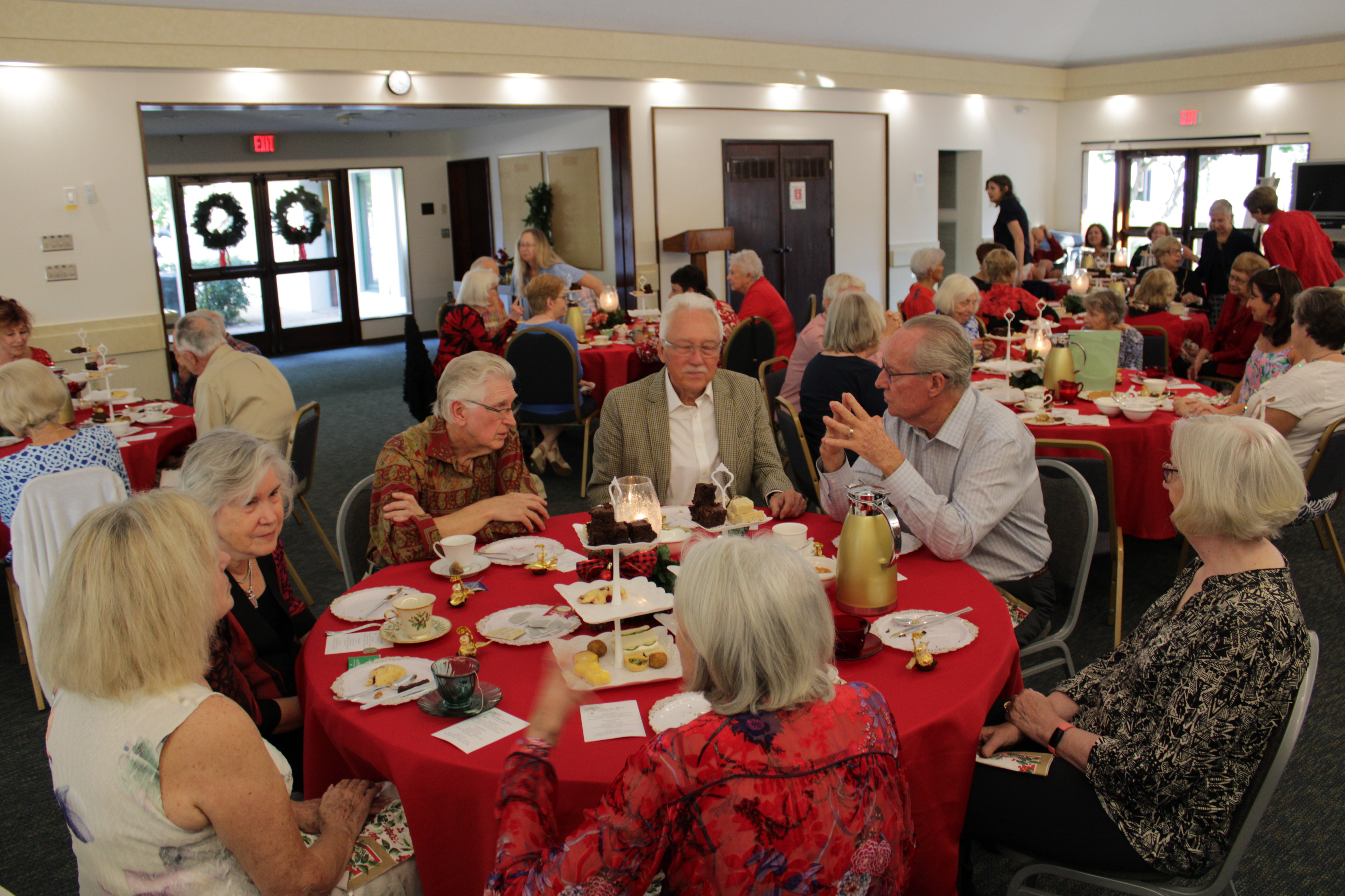 A group of people gathered around tables decorated with red tablecloths, enjoying refreshments and engaging in conversation during a community event.