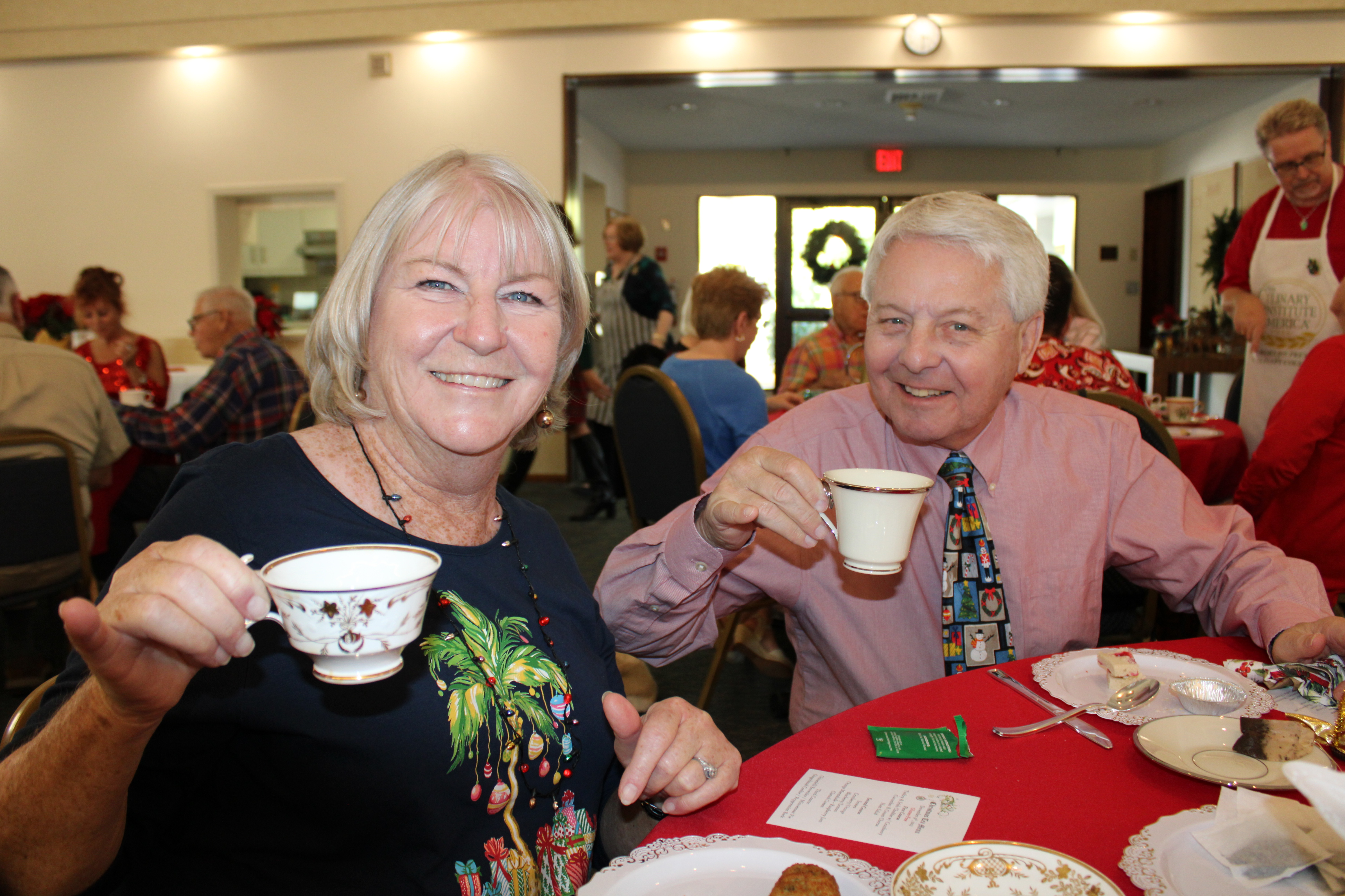 A joyful gathering featuring two smiling individuals holding teacups at a festive event, surrounded by a cheerful atmosphere and decorations.