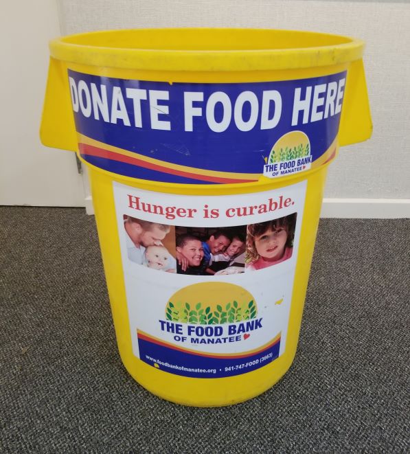 Yellow donation bin with a sign that reads 'DONATE FOOD HERE' and the message 'Hunger is curable.' featuring images of families and children, promoting the Food Bank of Manatee.